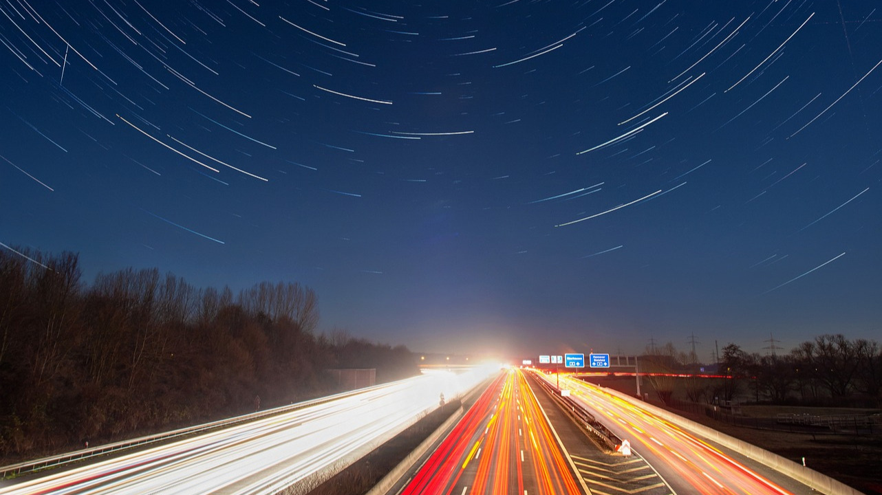 Vue en time-lapse d'une autoroute de nuit, avec des traînées lumineuses et un ciel étoilé en rotation, symbolisant l'accélération technologique.